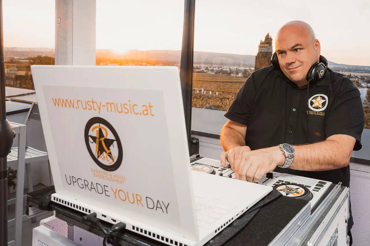 Hochzeits-DJ Rusty mit Kopfhörern am weißen DJ-Controller bei Sonnenuntergang, davor der weiße Laptop mit UPGRADE YOUR DAY