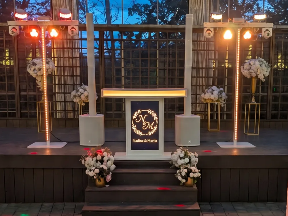 Konzeptbild des weißen Signature-Podiums mit Monogramm auf dem Bildschirm, daneben zwei weiße Säulen-PAs und Lichtstative im Glashaus