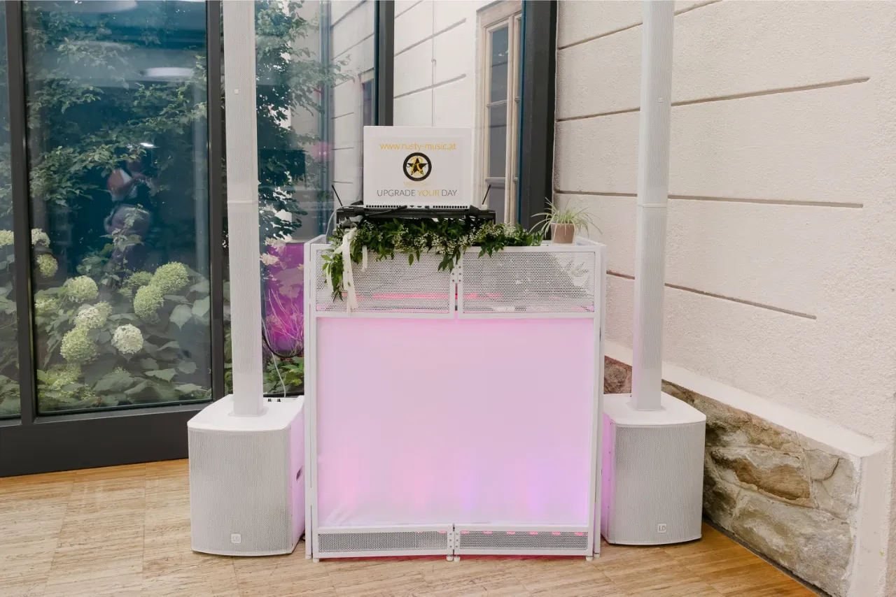 Weißes Hochzeits-DJ-Setup mit Säulen-PA und hinterleuchtetem DJ-Tisch, geschmückt mit Grün und Blumen