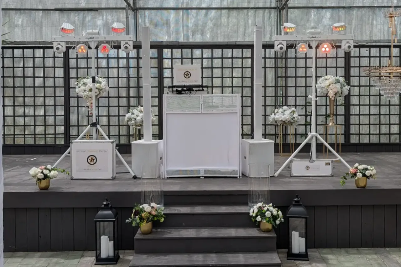 Weißes DJ-Setup mit Säulen-PA, DJ-Tisch und zwei Lichtanlagen auf einer Bühne im Glashaus, geschmückt mit weißen Blumen und Laternen